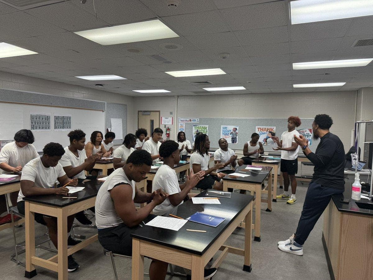 Young Black men in white shirts sit at lab tables, listening to a man speaking.