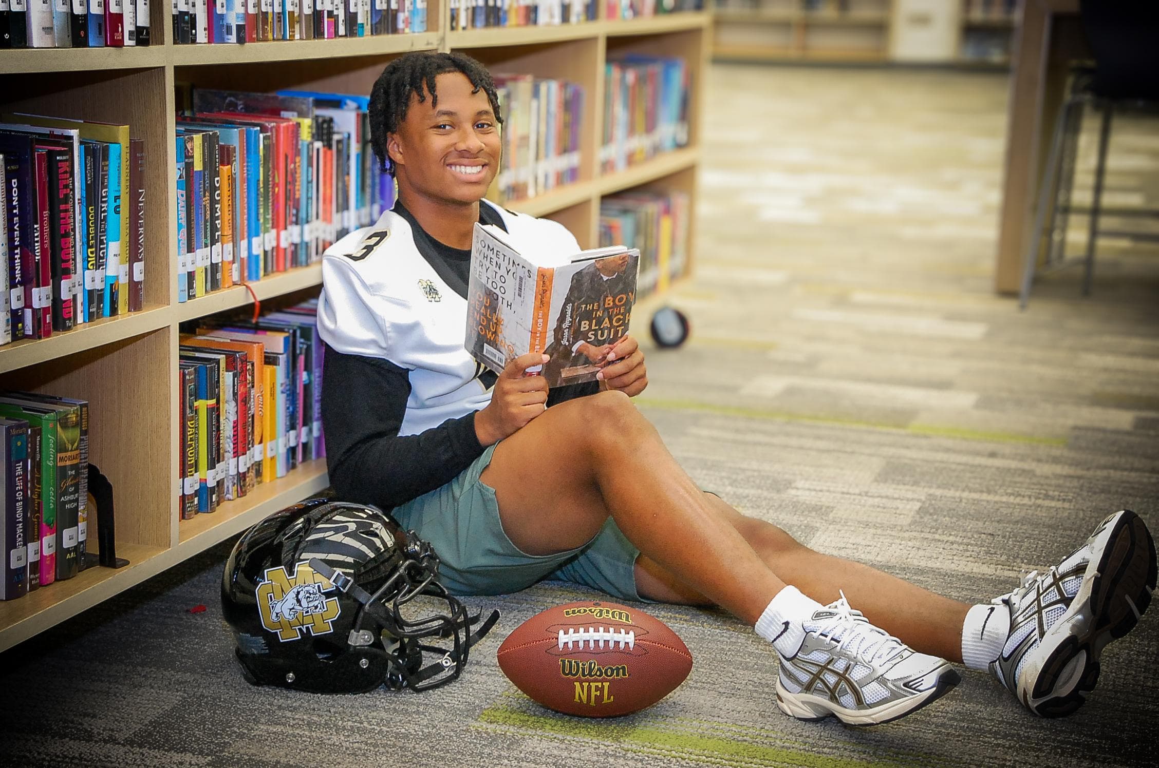 Smiling student-athlete sits on a library floor reading a book with his football and helmet.