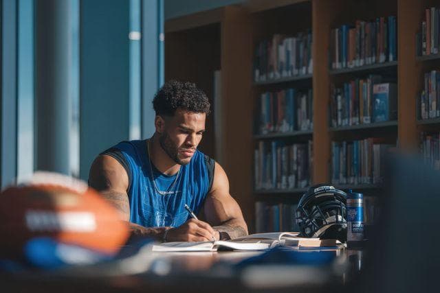 Muscular man in blue tank top writing at a library table with football gear.