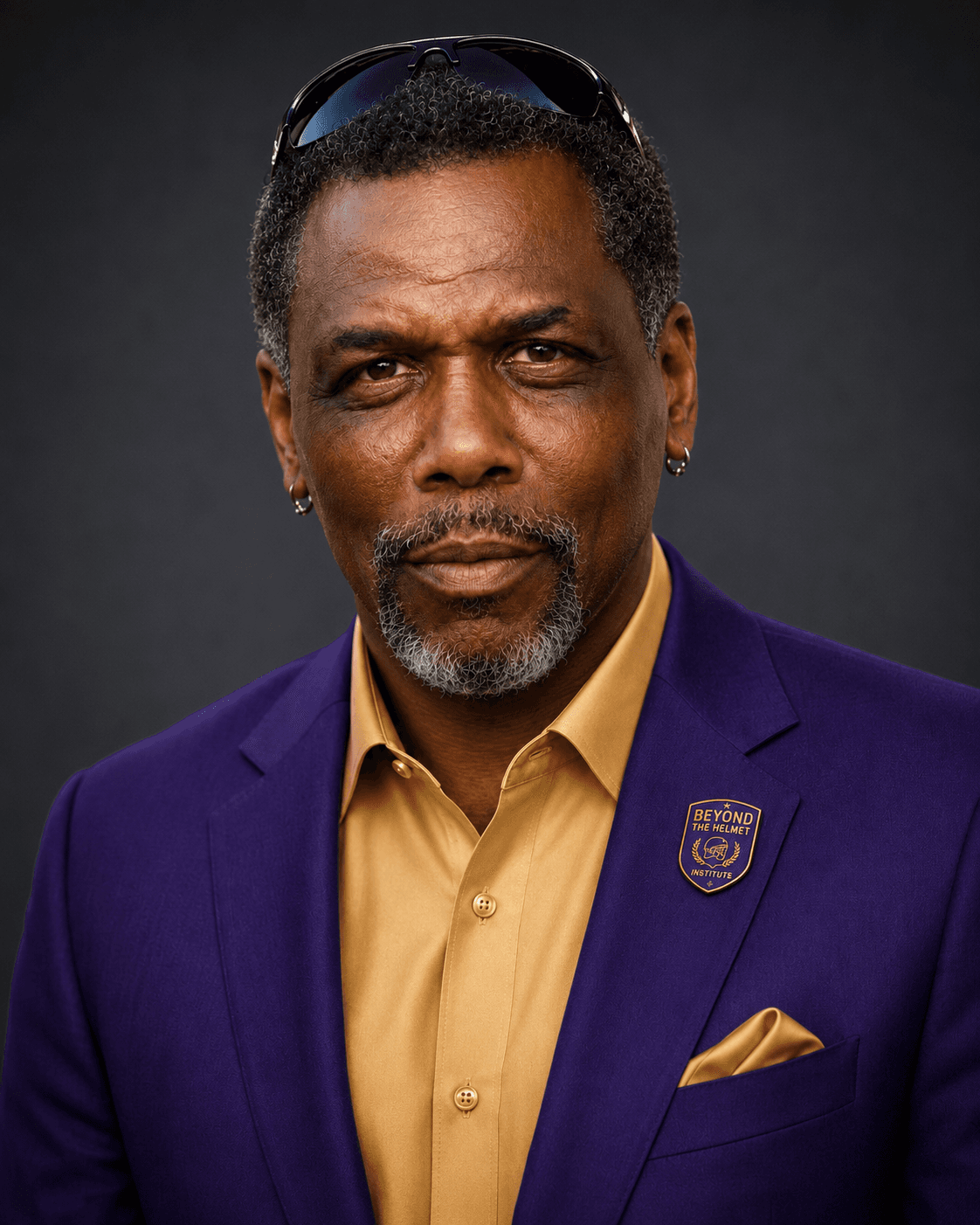 Middle-aged Black man with a goatee wearing a purple blazer, gold shirt, and lapel pin.