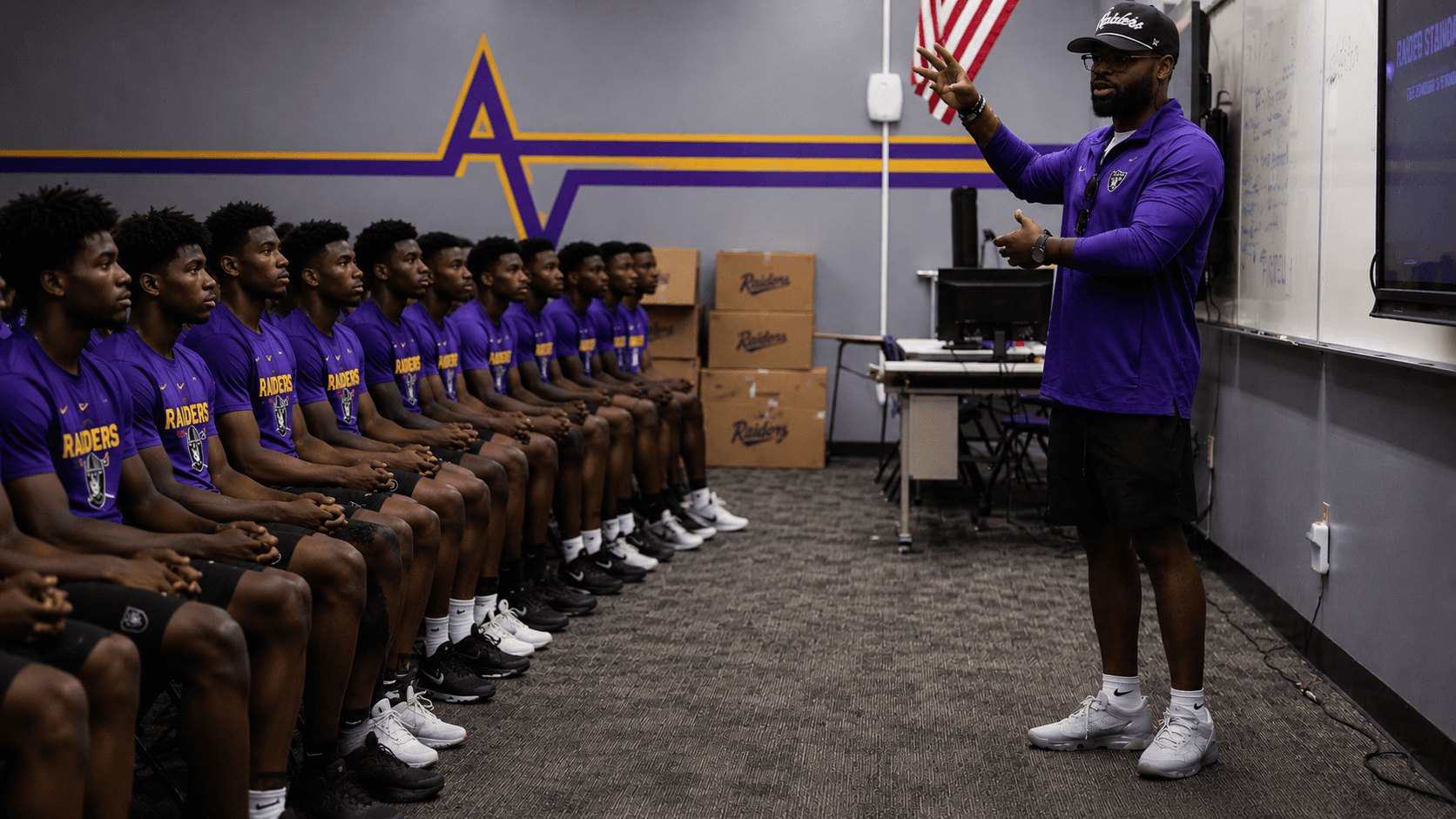 A coach gestures while addressing a row of seated athletes in purple Raiders shirts.