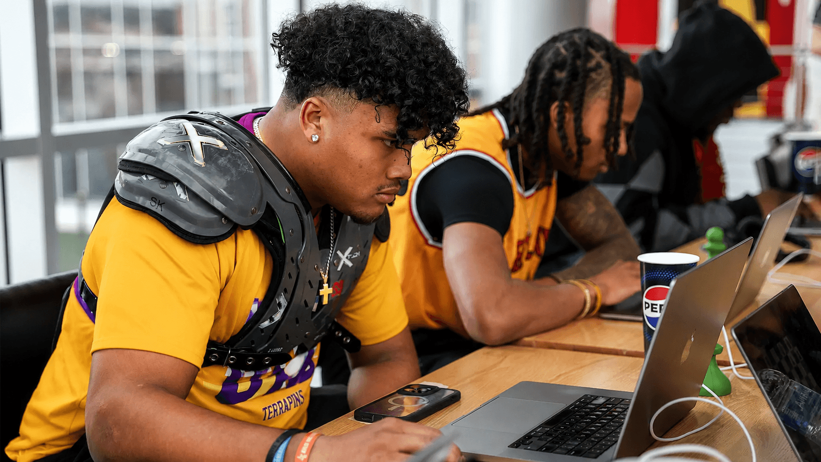 A Terrapins football player in shoulder pads and yellow shirt uses a laptop computer.